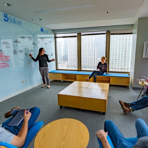 Image of a woman writing on a whiteboard, standing in front of a group of people taking notes.