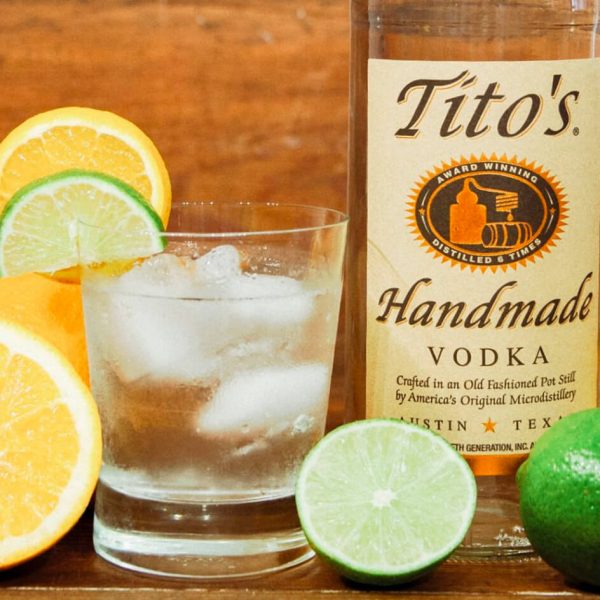 Image of a glass of vodka with ice in it, surrounded by slices of lemons and limes, next to a bottle of Tito's Vodka.
