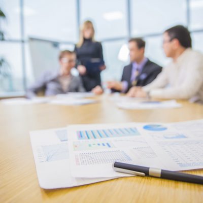 Image of a pen resting on top of papers with charts and graphs on them in the foreground, with people talking in the background.