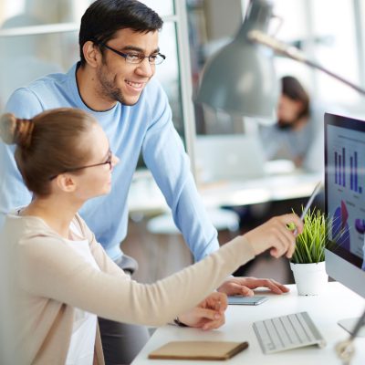 Image of a woman sitting at a desk pointing at her computer with a pen while a man stands next to her looking at her computer screen.